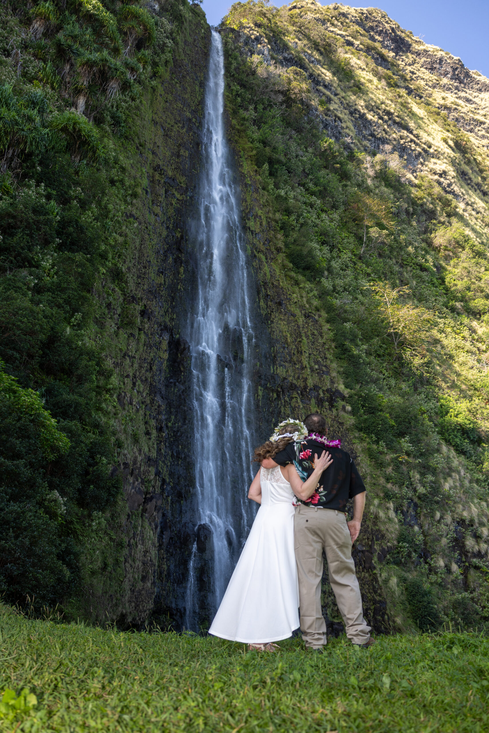 A couple in wedding clothes stands near a private waterfall on the big island of Hawaii
