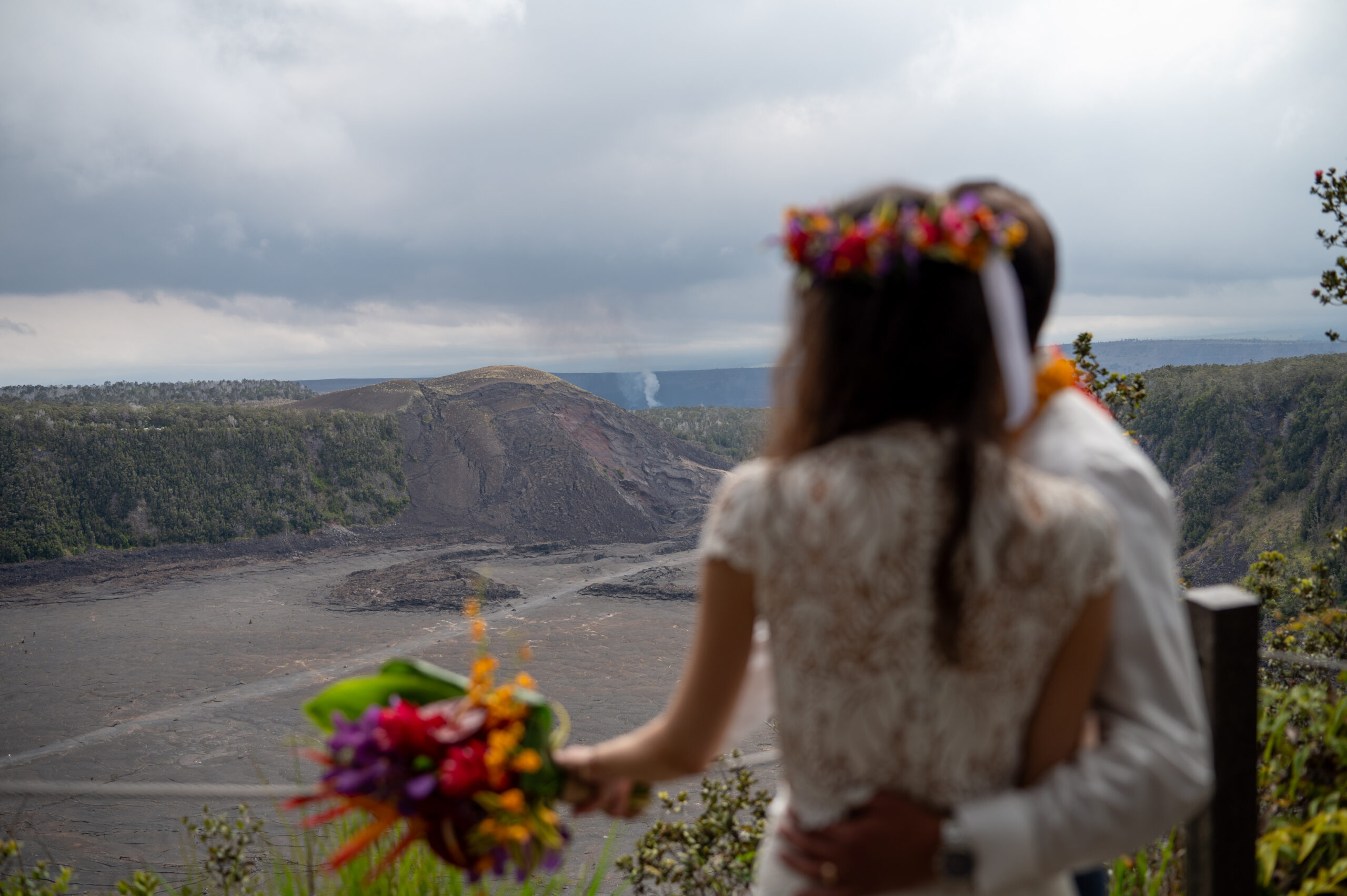 A couple in wedding clothes stands looking over Kilauea as it steams