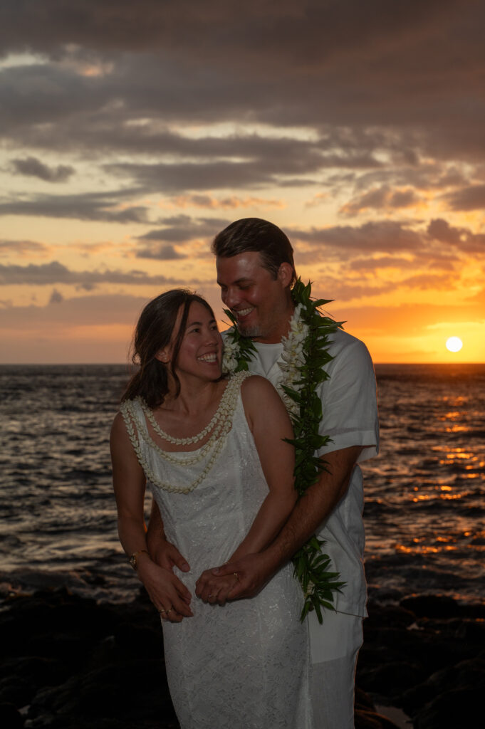 A couple in wedding clothes stands together in front of the sunset wearing traditional Hawaiian leis.