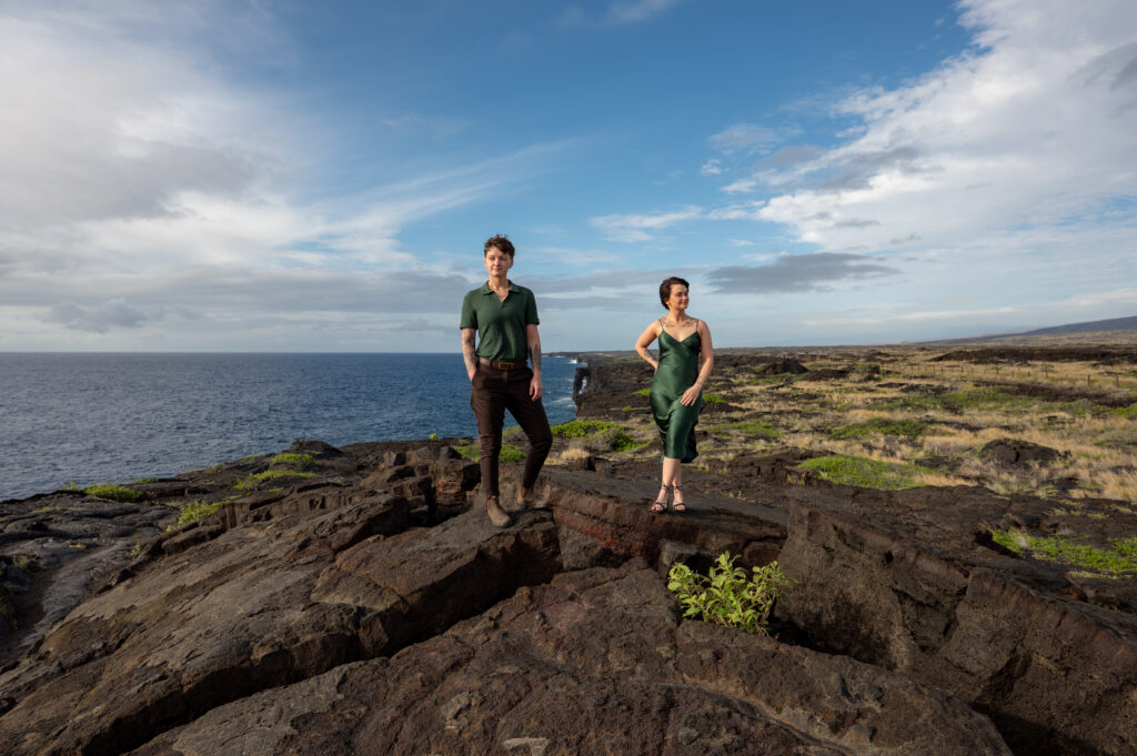 A couple stands on a lava rock formation with the Holei Sea Arch behind them