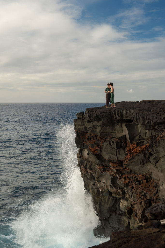 A couple stands near the edge of a cliff as a wave crashes into the cliff.