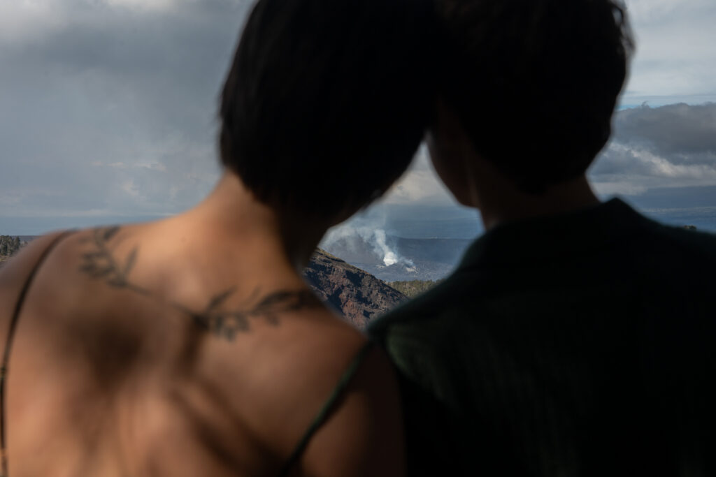 A couple stands with their heads together and between them is steam rising from Kilauea volcano in the distance.