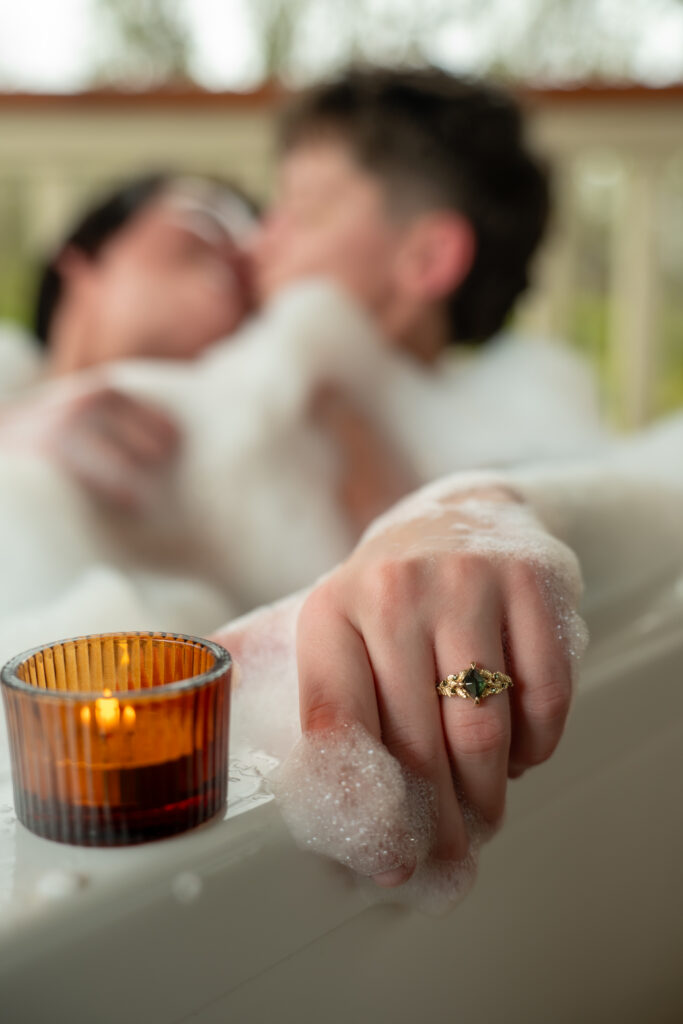 A couple sits in a bubble bath surrounded by candles on a lanai in Hawaii. The focus is on a green stone engagement ring.
