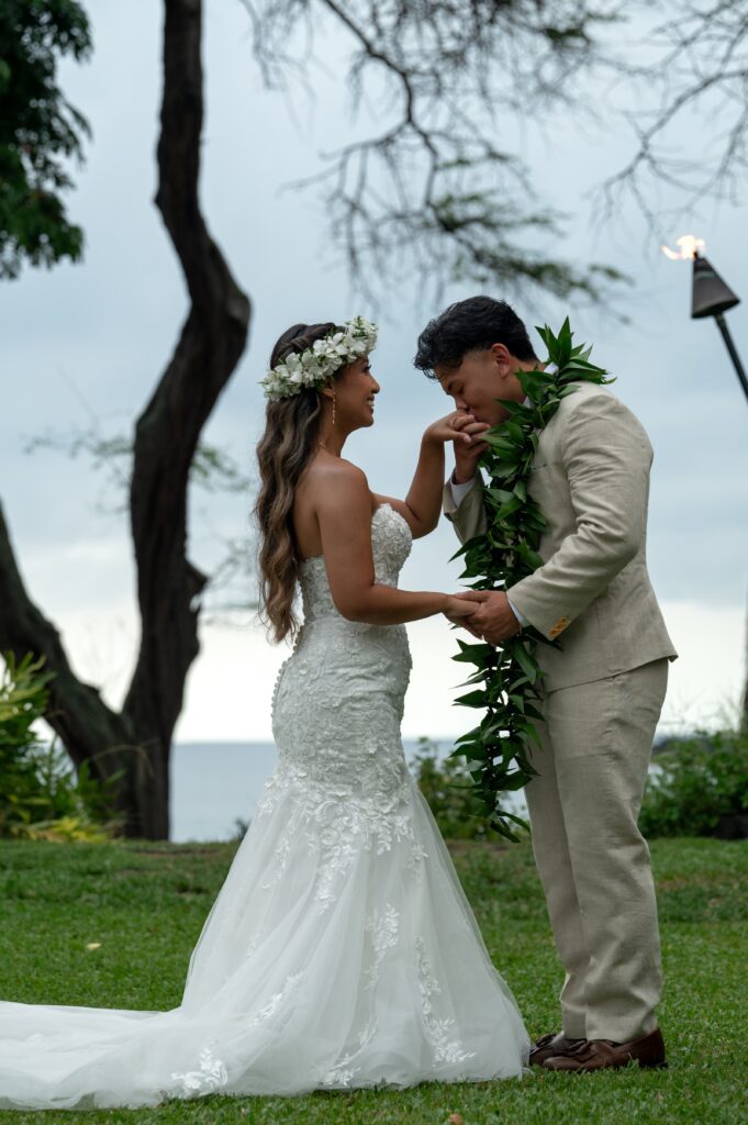 A groom kisses a bride's hand during their Outrigger Kona wedding ceremony on the Bayside chapel lawn